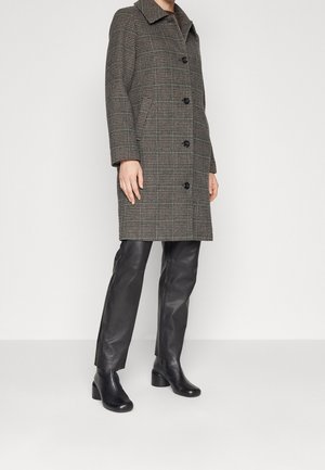 Osoba ubrana w kraciasty płaszcz do kolan z czarnymi guzikami, czarne skórzane spodnie i czarne botki na obcasie, stojąca na jednolitym tle.