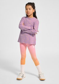Lila Langarmhemd mit gelbem Dreiecks-Muster, kombiniert mit ombre-farbenen Leggings in Pink und Orange mit weißen Chevron-Akzenten. Weiße Sneakers und Socken.