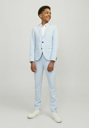 Completo azzurro chiaro con giacca a bottoni singoli, pantaloni slim-fit e camicia bianca. Scarpe da ginnastica bianche completano l'outfit. Tessuto morbido, design su misura.