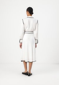 Robe blanche avec une silhouette A-line fluide, piping noir le long du col et des poignets, manches longues et taille ceinturée. Chaussures noires avec des accents.