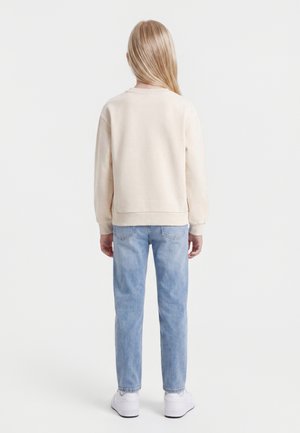 Enfant aux longs cheveux blonds portant un sweat crème, un jean bleu clair et des baskets blanches, debout de dos sur un fond uni.