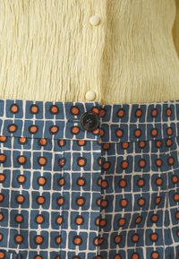 Žlutý texturovaný top s malými knoflíky, doplněný vzorovanou modrou látkou s oranžovými kruhy a mřížkovými liniemi, zajištěný černým knoflíkem.