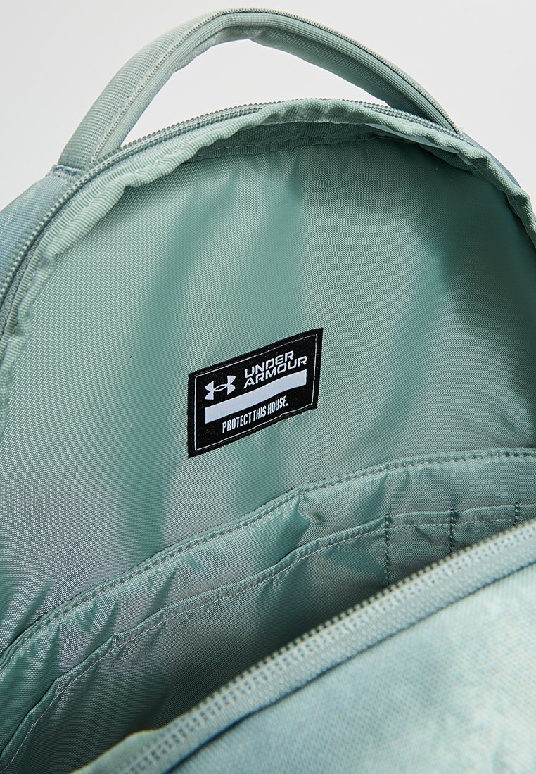 Interno dello zaino verde chiaro con tessuto liscio, etichettato "UNDER ARMOUR" e una targhetta nera con scritto "PROTECT THIS HOUSE", dotato di tasche.