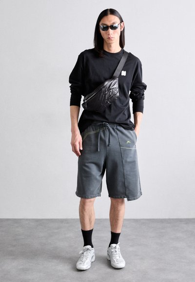 Schwarzes Langarmshirt, graue Shorts mit gelben Akzenten, schwarze Bauchtasche, schwarze Sonnenbrille, weiße Sneakers, mit einem eleganten, lässigen Design.