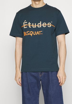 Man draagt een marineblauw T-shirt met grote beige tekst "Études" die doorgehaald is in oranje en daaronder oranje tekst "B. Squiat", gecombineerd met een blauwe spijkerbroek.
