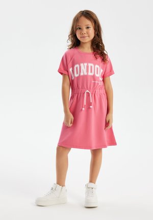 Rosa bomullsklänning med korta ärmar och dragsnörning i midjan. Har "LONDON" tryckt i vitt på framsidan. Matchad med vita sneakers.