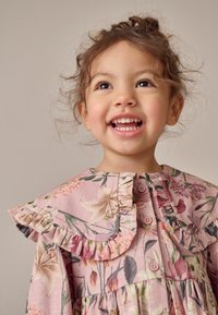 Květinové šaty z růžové látky s volánovým límečkem a zapínáním na knoflíky. Obsahují pastelové květiny a listy v různých odstínech.