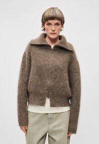 Sweater castanho, felpudo, com fecho de correr e gola alta, de corte curto e punhos canelados. Usado sobre uma camiseta de cor clara e jeans claros.