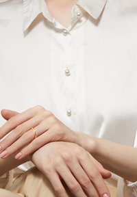 Gold ring with a wavy design, featuring small clear stones. Hands are resting on a light-colored fabric with a button-up shirt and pearl buttons.