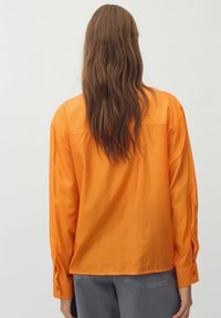 Camicia arancione a maniche lunghe con vestibilità rilassata, caratterizzata da polsini con bottoni e un tessuto liscio e leggero. Vista da dietro.