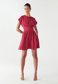 Czerwona krótka sukienka z dopasowaną talią i luźnymi rękawami, o gładkiej fakturze i rozkloszowanej spódnicy. Noszona z cielistymi sandałami na obcasach.