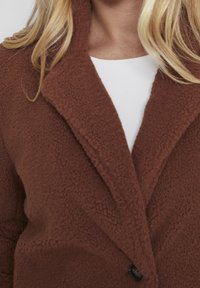 Brun fleecejakke med en struktureret finish, hakket krave og en enkelt sort knap. Har en afslappet pasform og et blødt udseende.