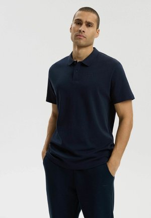 Mężczyzna o krótkich włosach, ubrany w granatową koszulkę polo z krótkim rękawem i pasujące spodnie, stojący z rękami w kieszeniach na jednolitym tle.