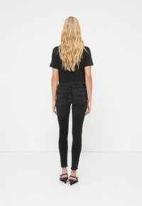 Femme aux longs cheveux blonds portant un t-shirt noir ajusté, un jean skinny noir et des sandales à talons noires, debout devant un fond blanc.