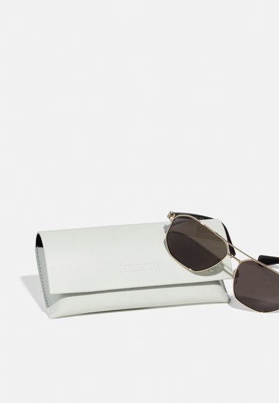 Óculos de sol com lentes pretas e armação dourada ao lado de uma estojo de couro cinza claro com o nome da marca em relevo "BURBERRY." Textura suave, forma alongada.
