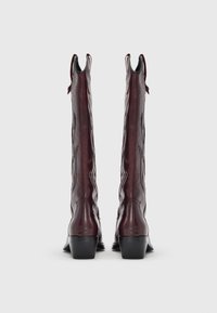 Ψηλές, σκουροκόκκινες δερμάτινες καουμπόικες μπότες με μαύρα κεντήματα και χονδρά τακούνια, εμφανίζονται από πίσω σε λευκό φόντο.