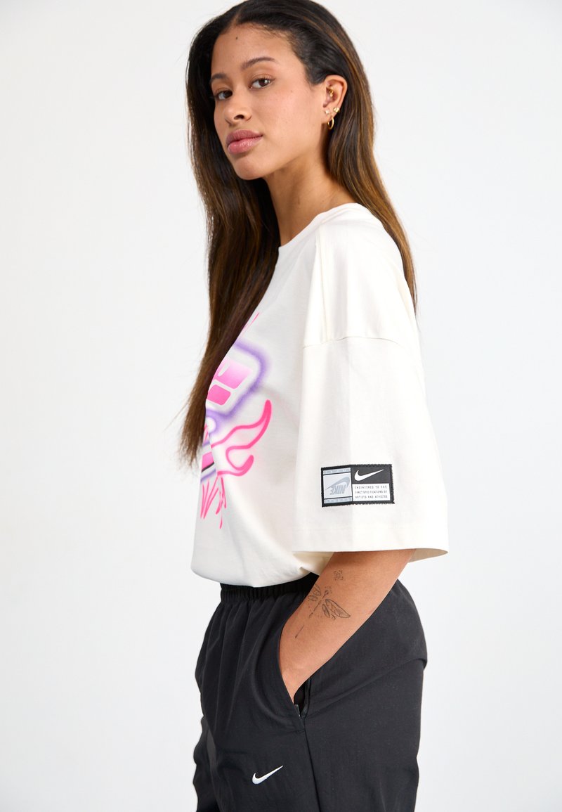 Weißes Baumwoll-T-Shirt mit oversize Passform, mit pinkem und lila Grafikdesign. Schwarzes Nike-Logo-Etikett am Ärmel. Schwarze Hose mit Taschen.