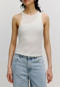 Weißes, geripptes Tanktop mit breiten Trägern und rundem Halsauschnitt, kombiniert mit hellblauen High-Waist-Jeans. Glatte Stoffoberfläche.