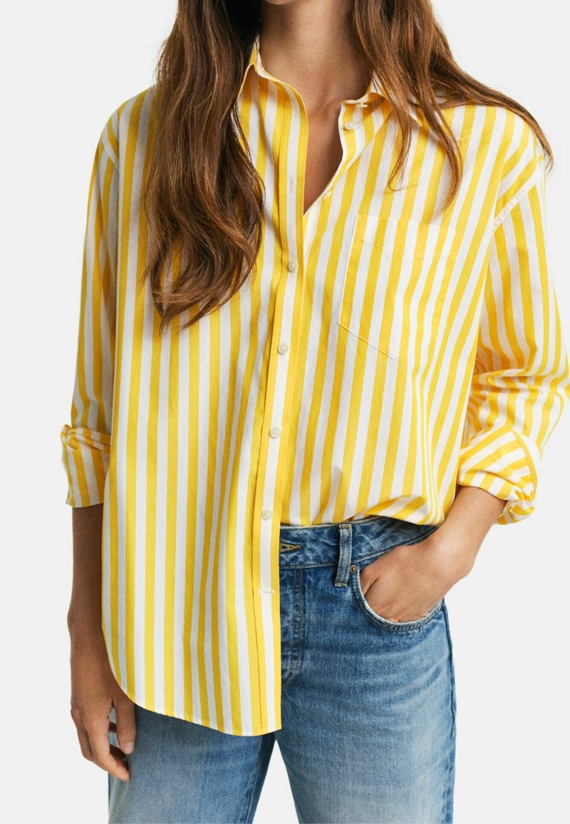 Camicia a righe gialle e bianche con bottoni, dotata di una tasca sul petto, maniche arrotolabili e una vestibilità comoda, abbinata a jeans blu.