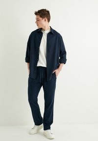 Navy-Button-Up-Hemd, darunter ein weißes T-Shirt, kombiniert mit navyblauen Jogginghosen und weißen Sneakers. Einfach, lässiger Schnitt.
