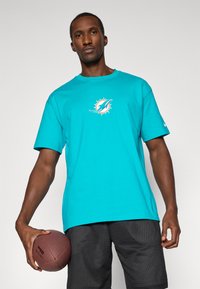Tyrkysové tričko Nike s logem Miami Dolphins na hrudi, v kombinaci se zvrásněnými černými šortkami. Model drží hnědý fotbal.