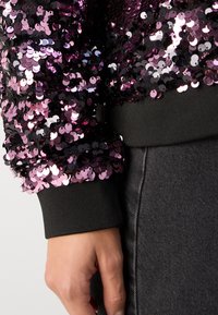 Prenda de lentejuelas negras y moradas con textura brillante, que presenta un puño negro acanalado y tela gris debajo. Primer plano de la manga.