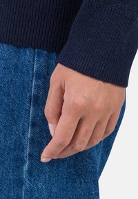 Der marineblaue Strickpulloverärmel liegt über einer blauen Jeans. Eine sichtbare Hand mit natürlichen Nägeln berührt sanft den Stoff und zeigt den Kontrast der Texturen.