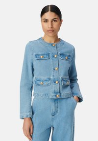 Jasnoniebieska kurtka dżinsowa ma okrągły dekolt, złote guziki, dwie kieszenie na piersiach oraz krótki krój z lekko wyblakłą teksturą.