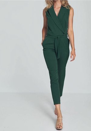 Mujer con un mono sin mangas de color verde oscuro con cinturón, pantalones ajustados y tacones beige con tiras caminando frente a un fondo claro y sencillo.