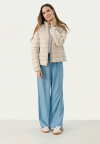 Giacca puffer beige chiaro con cuciture orizzontali, dotata di colletto rialzato e chiusura con zip, abbinata a pantaloni a gamba larga blu chiaro e scarpe bianche.