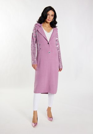 Lange roze jas met met pailletten versierde mouwen, knoopsluiting en een kraag. Draagt men over een witte top en gecombineerd met roze hoge hakken.