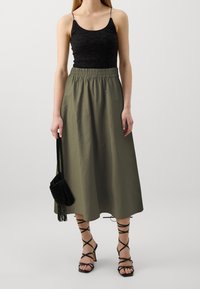 Schwarzes Spitzen-Camisole mit dünnen Trägern, olivgrüner, knöchellanger Rock mit elastischem Bund, kombiniert mit schwarzen Riemchen-High Heels und Tasche.