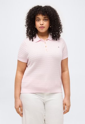 Femeie cu păr creț, purtând un tricou polo cu mâneci scurte, texturat, de culoare roz deschis și pantaloni albi, stând în fața unui fundal alb.