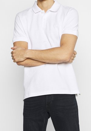 Homme portant un polo blanc à manches courtes et un jean noir, les bras croisés sur la poitrine, sur un fond uni.