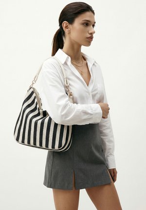 Donna che indossa una camicia bianca con bottoni e una gonna grigia, che porta una borsa a tracolla a righe bianche e nere con manico intrecciato.