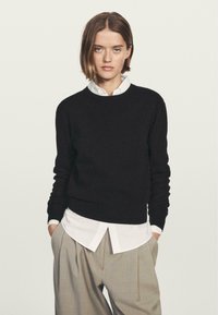 Maglione nero in tessuto morbido con scollo tondo, abbinato a una camicia bianca con colletto. Pantaloni grigi chiari a pieghe completano l'outfit.