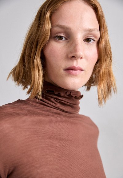 Barna bordás turtleneck felső fodros nyakkivágással, testhez simuló fazonnal és puha textúrával. Rövid, hullámos haj látható.