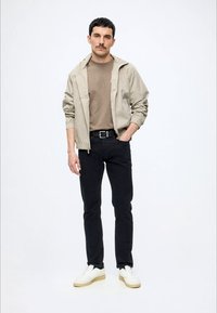 Mann steht, trägt eine beige Jacke über einem braunen Hemd, schwarze Hose mit schwarzem Gürtel und weiße Sneakers vor schlichtem weißem Hintergrund.