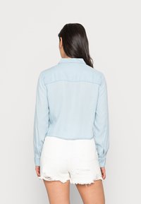 Camicia di jeans azzurro chiaro corta con maniche lunghe, colletto e orlo leggermente sfrangiato, indossata sopra pantaloni corti bianchi strappati.