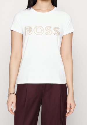 Frau trägt ein weißes T-Shirt mit kurzen Ärmeln und dem Wort BOSS in großen beigen Buchstaben und dunkelroten Hosen.