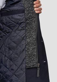 Gewatteerd marineblauw jack met grijze gebreide accenten, voorzien van een zijrits, gevoerde binnenkant en een gladde, gestructureerde stof.