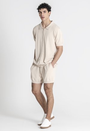 Joven de pie con las manos en los bolsillos, vistiendo una camisa beige de manga corta con cremallera, pantalones cortos a juego y zapatillas blancas sobre un fondo liso.