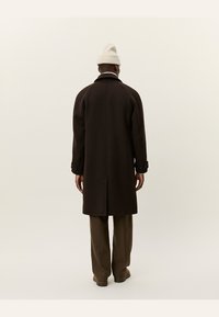 Brown wool coat with a tailored fit, raglan sleeves, and buttoned cuffs, paired with light-colored trousers and a knitted hat.