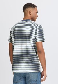 Trumpas rankovės juostuotas marškinėliai su tamsiai mėlynomis ir baltomis horizontaliomis juostelėmis, apvaliu kaklu, pagaminti iš medvilnės, laisvas kirpimas. Matomos mėlynos džinsai.