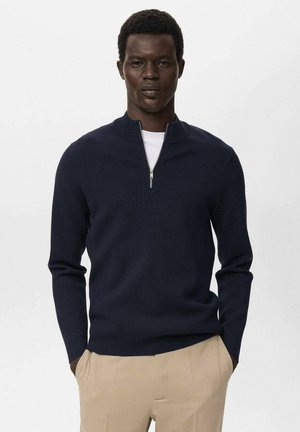 Suéter de punto azul marino con alto cuello y cierre de media cremallera. Realizado en una tela suave, de diseño ajustado y de mangas largas. Combinado con pantalones beige.