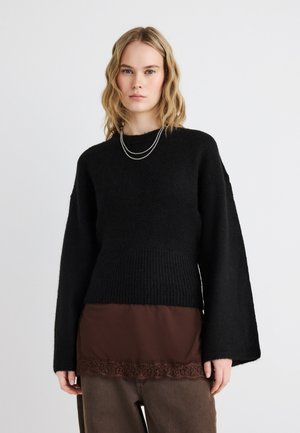 Tânără cu păr blond ondulat, purtând un pulover negru cu mânecă lungă, peste un top maro cu dantelă și pantaloni maro.