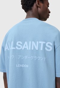 Vaaleansininen suuri T-paita, joka on valmistettu puuvillasta, ja jossa on rohkeaa valkoista tekstiä selässä: "ALLSAINTS" sekä lisämerkkejä ja "LONDON."