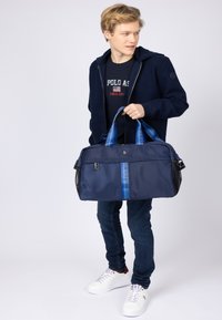 Námořní duffel taška vyrobená z nylonu, s modrými popruhy s textem, zipovým uzávěrem, bočními síťovými akcenty a logem na přední straně.