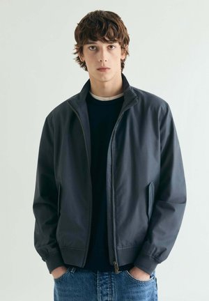 Joven con cabello castaño, vestido con una chaqueta de cremallera azul marino oscura y jeans azules, de pie con las manos en los bolsillos contra un fondo claro y liso.