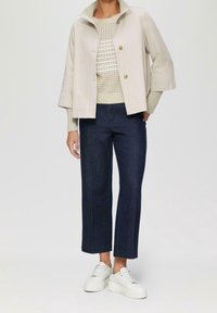 Persoon in een beige cropped jack over een crèmekleurige gebreide trui, donkerblauwe rechtopstaande jeans en witte platform sneakers, staande tegen een effen achtergrond.